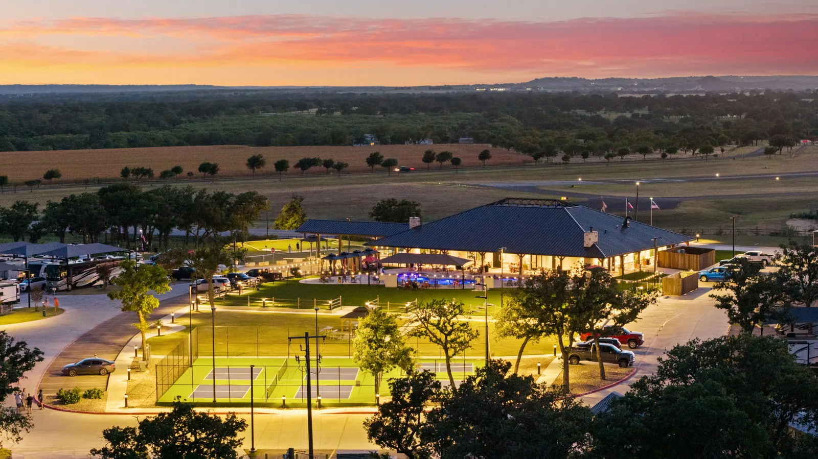 Lantern Oaks RV Resort at dusk, with the lit clubhouse, pool and pickleball courts among live oaks and Hill Country ridges beyond